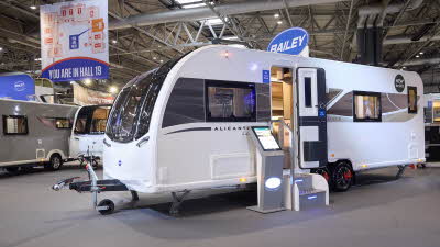 Bailey Alicanto Grande Deluxe Alvor’s exterior is white with brown decals from the front to the rear.  The entrance door is open and there are two metal steps to gain easy access.  There is an interactive stand to the left.