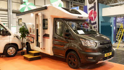 The Pilote Atlas A690GJ has a black cab and a white coachbuilt body which is decorated with black and red decals.  The habitation door is open and there are two steps to gain easy access.  There is an information stand and a plant to the left.