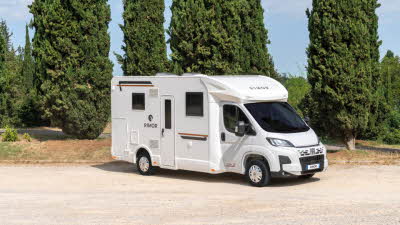 The Rimor Sarus 67 Plus has a white exterior with gold and black decals.  It is parked in front of five pine trees