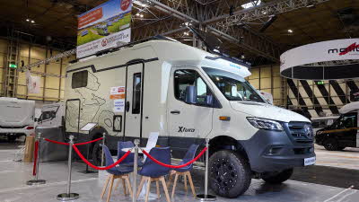 The Eura Mobil Xtura XT 686 EF is cream with black trim and decals.  The entrance is sealed by stanchions carrying red rope.   There is a table outside with four blue chairs.  