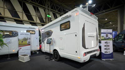 Auto-Trail Excel 690T’s exterior is white with dark grey decals.  It has a large door to the rear for its garage.  There is an interactive stand next to the rear wheel.  There is a roller banner at the end of the vehicle.