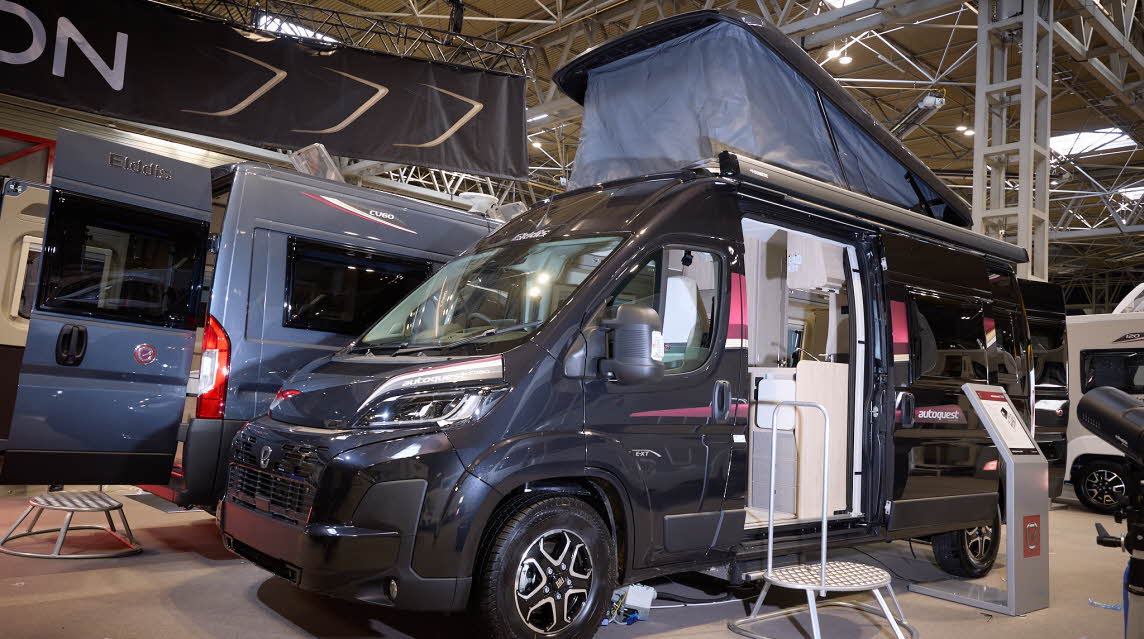 The Elddis Autoquest CV80 is black.  There is a metal step in front of the sliding habitation door which is open revealing the interior.  There is an interactive stand to the right.  The pop top roof is fully extended.