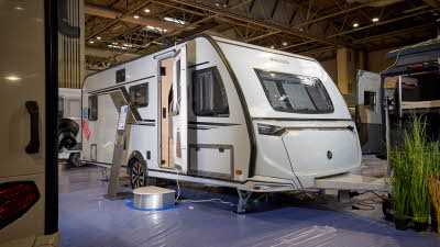 The Knaus Südwind 580 QS Black Selection is white with black edging, there are plants by its hitch.  The entrance door is open and there is a metal step to gain easy access.  There is a stand next to the steps.