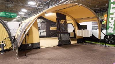 The Raclet Safari GL has a dark cream coloured canvas.  The door is open revealing the interior.  There is a round table covered by a tablecloth in the covered outside area.  