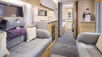 The interior of the Adria Adora 612 DL Seine has cream walls.  The furniture is mid wood.  There are two sofas with grey upholstery and on one there is a head rest consisting of a grey and a purple cushion.  The washroom is at the rear.