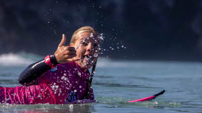 A young surfer lying down on their surfboard on the water looking at the camera giving a thumbs up as the water splashing gently with the movement
