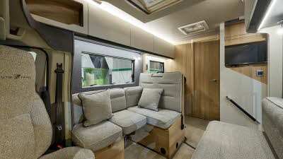 Auto-Trail Excel 675B’s interior has beige fabric upholstery with matching cushions.  The furniture is wooden.  There is a TV above the handrail by the entrance door.  There is one sky light.  The internal door to the washroom is closed.
