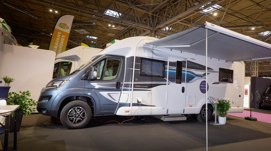 The Coachbuilt GB LIV Swift Escape 674 has a great cab with white body.  There are grey and turquoise decals.  There is a large canopy awning with a stand and plant to the right of the habitation door.