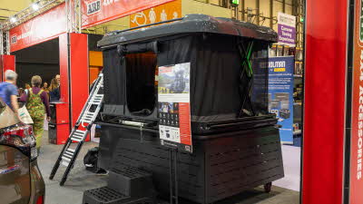 The ARB Altitude has black canvas and a grey hard shell roof.  The front door is open.  It is displayed on a stand and there are two steps to gain easy access.  The construction struts are green.