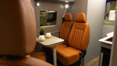 The AURA 6EB's upholstery is orange leather.  The table has two mugs and a pot plant on it.  Toward the rear is the bed with a green cover on it.