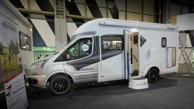 Auto-Trail Excel 675B’s exterior is white with a silver cab and dark grey decals.  Its entrance door is open revealing a handrail and a blue umbrella.  There is a step to gain easy access.