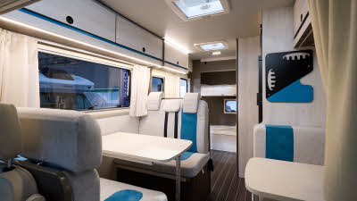 Rimor Kilig 9’s interior has cream upholstery with turquoise inserts.  Its furniture is pale wood again with turquoise inserts.  It has a wood effect floor.  There are two separate tables.  There's two fixed bunks at the rear.