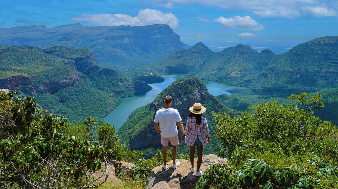 Two people standing on rocks looking over the stunning the Blyde river canyon with lush greens blue river and pale blue sky with fluffy clouds