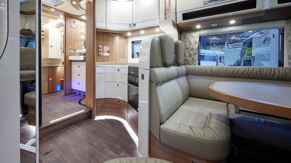 Carthago chic e-line I 64 XL QB MB has leather upholstery with curved cream overhead lockers with wooden surrounds.  There is a large oval table.  There is a wooden floor.  The kitchen has curved white storage lockers and a full oven.