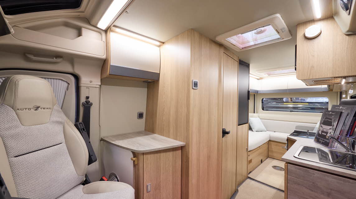 The Auto-Trail V-Line 635 Sport has beige carpets, with pale wood cabinets with cream and grey overhead lockers.  The upholstery is white and cream.  There is a rear lounge.  The front seats are swivelled around.  There is a roof light.