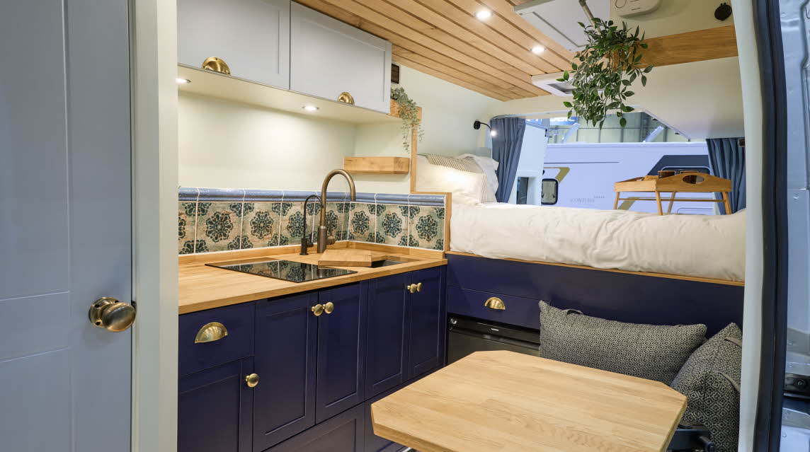 Vanlife Conversions Kunu has wooden flooring with grey upholstery, dark blue units with gold handles.  There is a fixed rear bed and the rear doors are open.