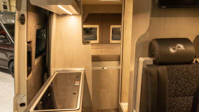 The Mega Mobil Mega Classic 600 S Hubbett has brown upholstery.  Its furniture is pale wood.  There is rear fixed bed with the rear panels complimenting the upholstery.  The kitchen is to the front left.