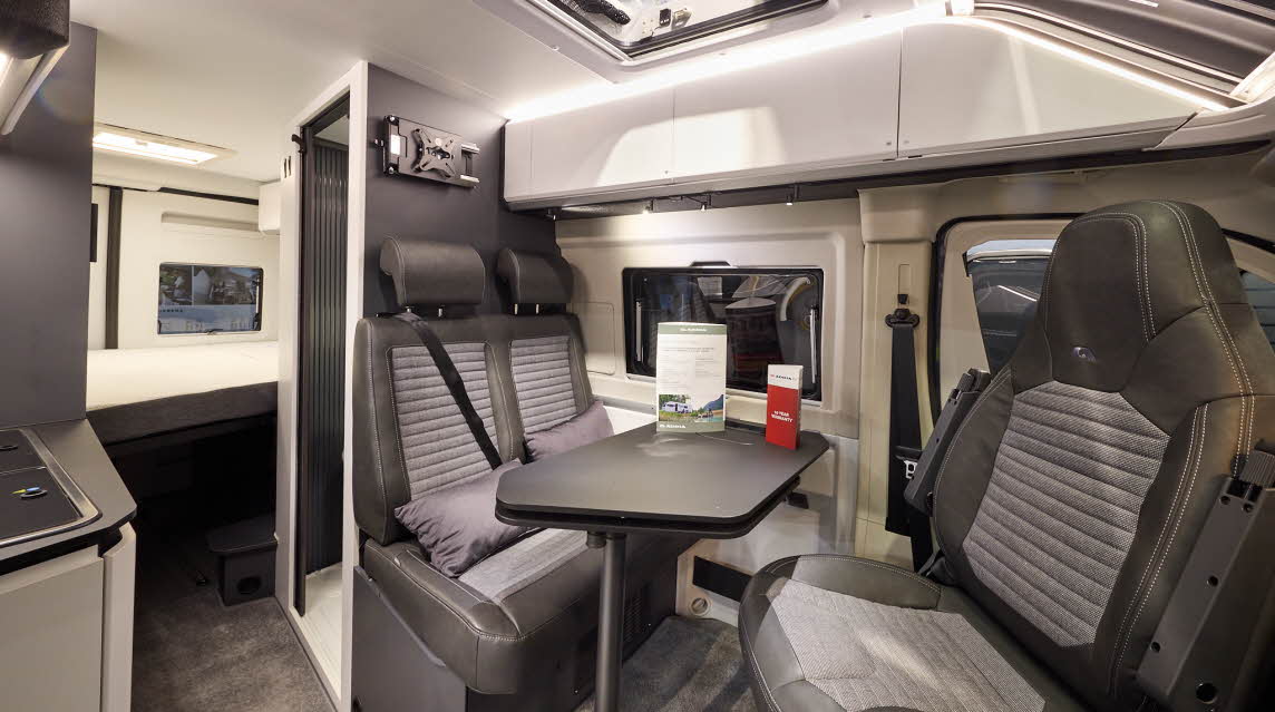 The Adria Twin Sport’s floor has a mid brown carpet, with white cabinets and dark grey panels.  Its rising roof is open.  The upholstery is two tone with dark edging.  There is a grey table which can be rotated.  