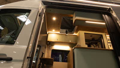 The Knights Custom Conversions Prestige XL has a wooden ceiling with a pattern, the cabinets are green with wooden edging and there is strip lighting underneath.  The door to the central washroom is open.