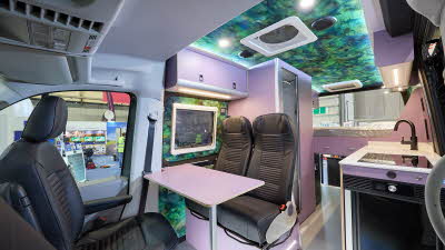 Vanworx MaxTraxx Crafter’s interior has black leather upholstery.  There is a white table and its furniture is white.  It has a patterned internal roof.  At the rear there is a fixed bed.