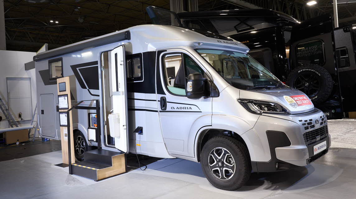The Adria Compact Supreme has a silver exterior.  The habitation door is open and there are two steps to gain easy access. There is an information board to the left of the steps.  