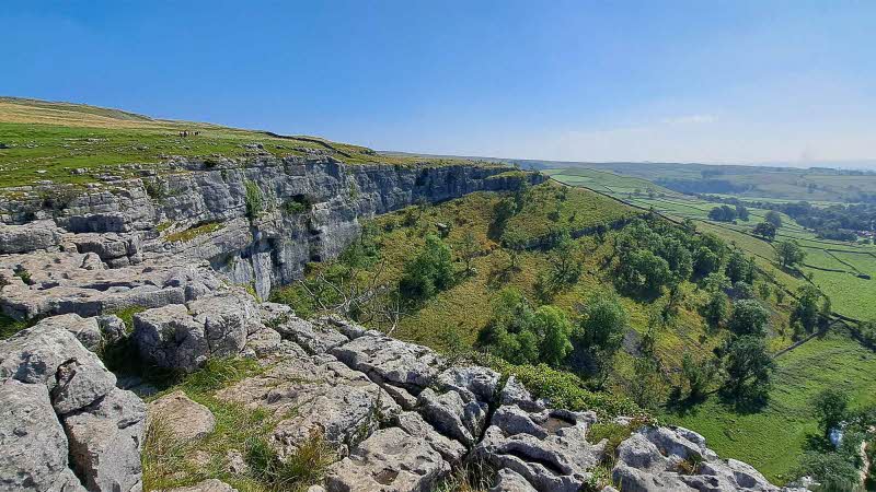 Standing at the top of Malham Cove, the rock face curving round into the distance with green grassy banks below, under a blue sky.