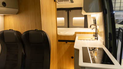 The Project Yonder Nomad 4X has black upholstery with green and cream locker.  The kitchen is to the right.  There is a rear fixed bed which a cupboard underneath.