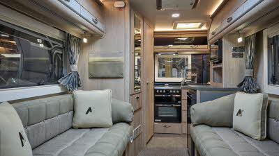 Auto-Sleeper Bourton's interior has two tone beige upholstery with cream and grey cushions.  There are tied cushions at the windows either side.  The wooden furniture has grey highlights.  There are two skylights and the kitchen is at the rear.