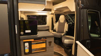 The VANTourer 630 L has brown and cream upholstery with matching cabinets.  The front two seats are swivelled and face the dining table and rear passengers.  The sliding door is open.