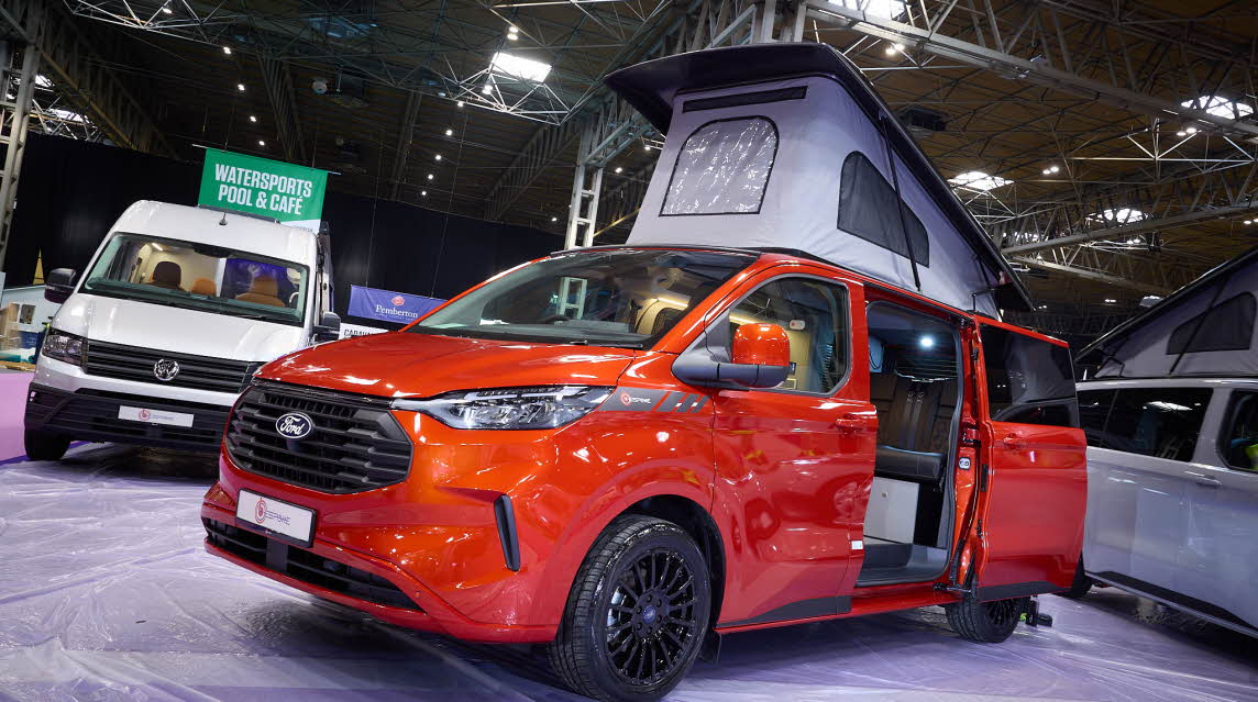 The Bespoke Campers Explore is orange with black trim.  The habitation sliding door is open which reveals the interior.  The light grey pop top roof is fully open.