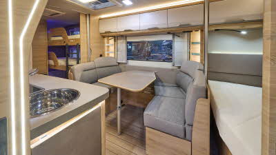 Interior of the Knaus Südwind 580 QS Black Selection with grey wraparound sofas, wooden accents and a wooden floor.  The fixed French transverse bed is at the front and the bunks are at the rear.