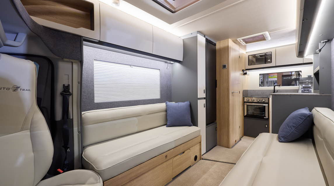 The Auto-Trail Excel 620S has beige carpet, with pale wood cabinets with white overhead lockers.  The upholstery is cream leather.  There are blue cushions.  The kitchen is at the end with an oven and a microwave in an overhead locker.