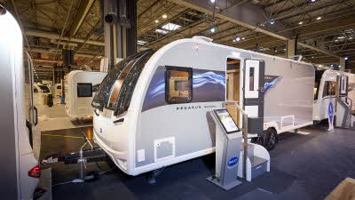 The Bailey Pegasus Black Bologna has a grey exterior with a grey decal including pale blue ripples on it,  The front door is open and there is an interactive stand next to the two metal steps.