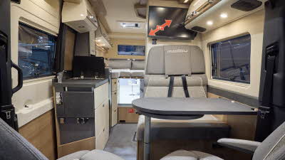 The Malibu Genius 641 LE has grey flooring with cream and wooden cabinets.  The upholstery is beige and grey.  There is a grey table in front of the sofas.  There is a step to the fixed rear bed.  The rear doors are open showing the storage.