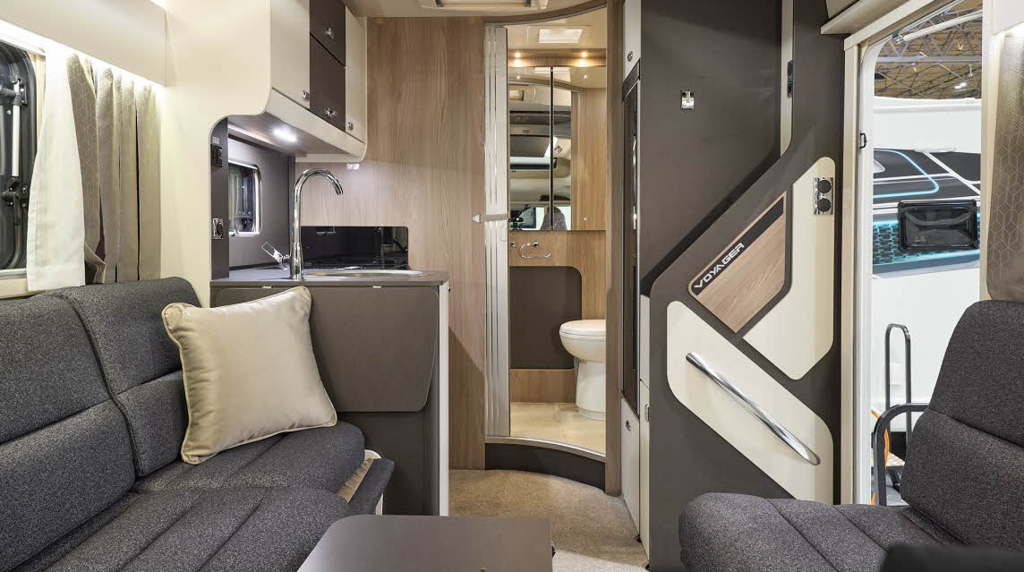 The Swift Voyager 540 has wooden flooring with dark grey upholstery, there is a gold cushion on the sofa.  The cabinets are dark brown and cream.  There is a step to the rear washroom.