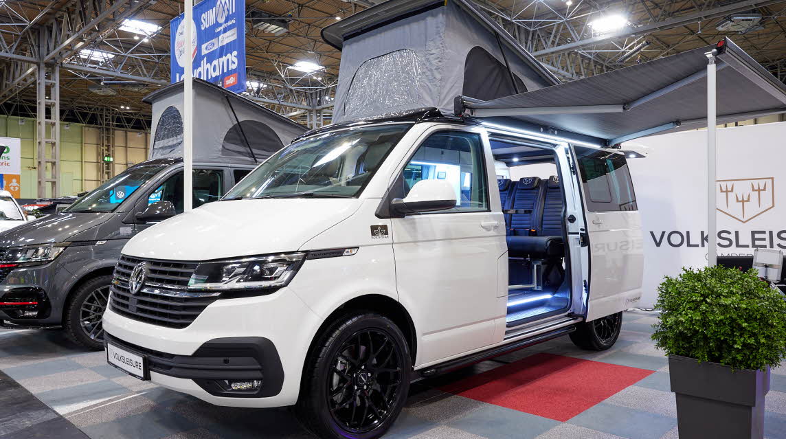 Volksleisure White Star has a white exterior.  The sliding door is open showing the interior, there is a strip light inset on one of the steps.  The rising roof is fully risen and the built in awning is extended.
