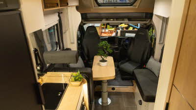 The Pilote Atlas A690GJ has black and grey upholstery.  The front seats are swivelled to the dining area where there is a wooden extendable table with a bonsai tree on top.  The floor has grey tiles. 