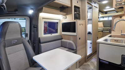 RP Motorhomes Rebellion 2 has two tone grey upholstery.  There are light wooden overhead lockers and door to the washroom.  On the floor is wooden laminate.  There is a folding flap which gives additional kitchen work space.