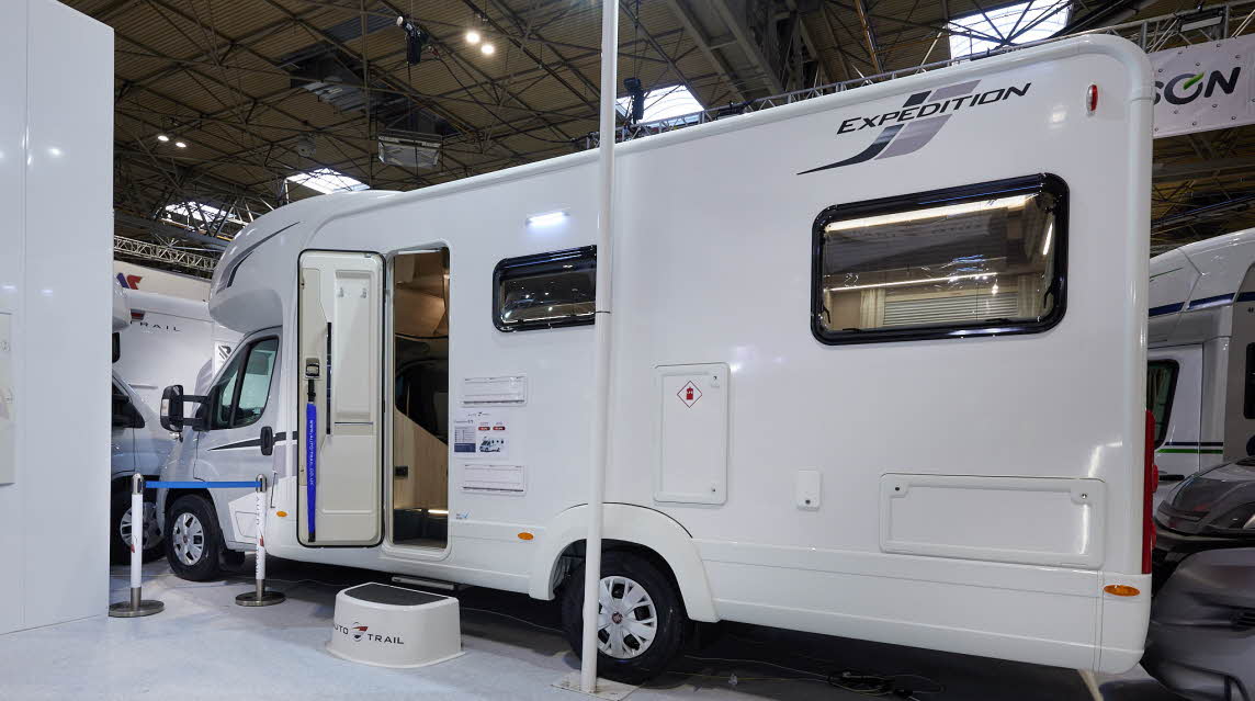 Auto-Trail Expedition C72 exterior, the motorhome is white with two windows, the habitation door is open showing into the interior, with a step to gain easy access.  There is a blue Auto-Trail umbrella in the door.