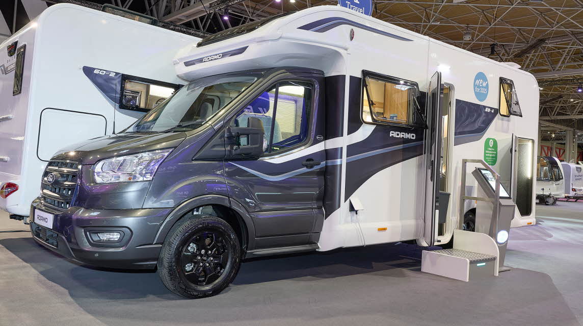 The Bailey Adamo 69-4 has a grey cab with a white body and has grey and blue decals.  There is a white and metal step by the habitation door.  There is an interactive stand to the right of the step. The rear garage door is open.
