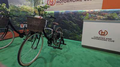 A bicycle with a wicker basket full of flowers is parked in front of a desk with the Caravan and Motorhome Club logo