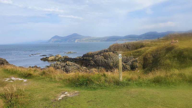 On the grassy edges looking out over the sea at Porthdinllaen on the Llyn Peninsula. The sky is pale blue with white clouds above the faded peaks across the waters  