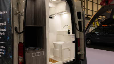 The Rolling Homes Camper Ltd Darwin FL (MWB)'s rear doors are open revealing the rear washroom which is white.  There is an orchard next to the wash basin.  Next to the washroom is a large storage area.