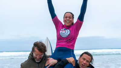 A surfer celebrating with arms in the air while being lifted up on the shoulders of two people