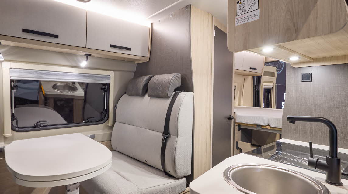 Xplore CV X9 has cream upholstery with brown headrests.  The cabinets are cream with wooden surrounds.  There is a round kitchen sink.  There is a fixed rear bed and the rear doors are open.