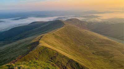 An elevated view from the top of a mountain ridge in the Brecon Beacons. The ridge and slopes disappearing in the distance in the low clouds under a orange sunlight fading sky