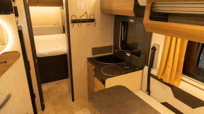 The Rimor Sarus Plus 66 has coffee and cream upholstery complemented by wooden furniture.  The kitchen is in the right corner.  To the rear there is a fixed bed in the bedroom.