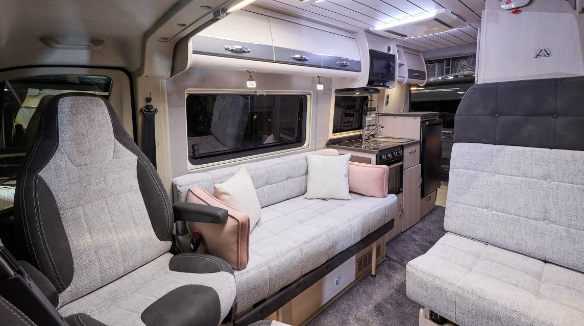 The Auto-Sleeper Symbol has grey carpet, with pale wood cabinets with pale grey highlights.  The upholstery is cream and dark grey.  There are cream and peach cushions.  The kitchen is in the middle and has an oven with a microwave overhead.