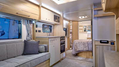 Bailey Pegasus Grande Bologna interior, it is grey with light wood.  There is a fixed bed beyond the kitchen with an end washroom, the door of which is open and is covering a large mirror.