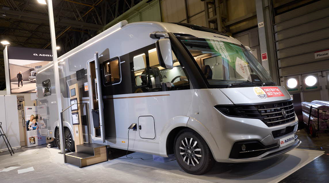 The Adria Supersonic has a silver exterior.  The habitation door is open and there are two steps to gain easy access. There is an information board to the left of the steps.  